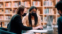 Twee vrouwen en een man met mondkapjes kijken op laptop in bibliotheek