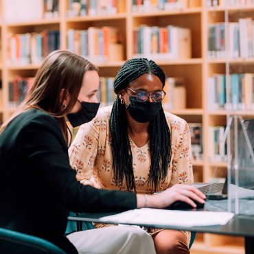 Twee vrouwen en een man met mondkapjes kijken op laptop in bibliotheek