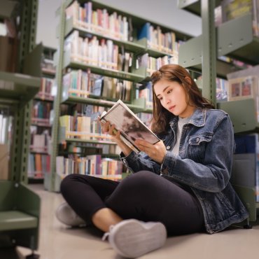 Jonge vrouw zit op de grond in een bibliotheek en leest een boek.
